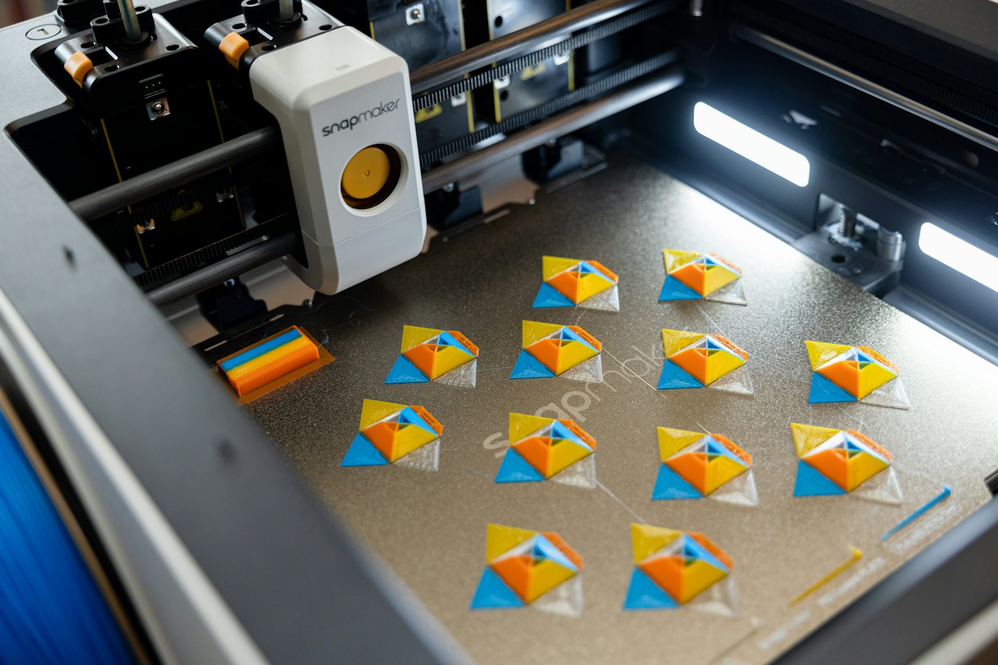 3D printer creating small blue, orange, and yellow geometric objects.
